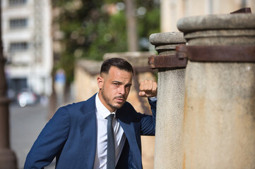 A young, handsome, dark-skinned Latin man wearing a suit and tie rests his fist on a stone column...