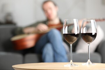 Glasses with red wine on table and man playing guitar indoors, selective focus