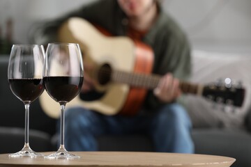 Glasses with red wine on table and man playing guitar indoors, selective focus
