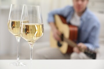 Glasses with white wine on table and man playing guitar indoors, selective focus
