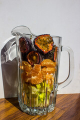 A glass jug, filled with various fruit pieces, including apple, Mandarin and Passion Fruit halves,...