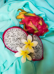 A sliced and whole dragon fruit, decorated with Frangipanni blossoms, on a turquoise background. 