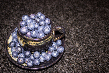 A black ceramic cup, filled to over spilling with large indigo-colored blueberries, with more blue...