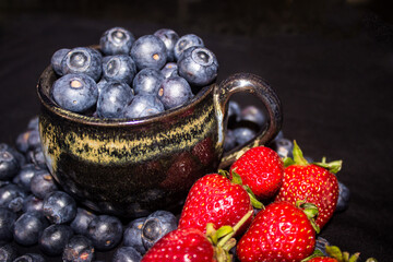 A black pottery teacup, filled to overflowing with blueberries, surrounded by strawberries and more...