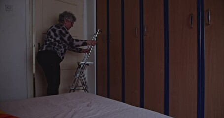 An elderly woman uses a stepladder to reach the top shelves of her closet. She is tidying and organizing her belongings.