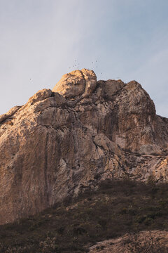 Pe&ntilde;a de Bernal, fotograf&iacute;a de una formaci&oacute;n rocosa producto de un volc&aacute;n extinto. Monta&ntilde;a al atardecer con una parvada de p&aacute;jaros sobrevolando la cima. 
