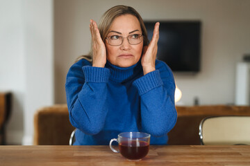 Woman expressing frustration while sitting at a table with a cup of tea in a cozy indoor setting...