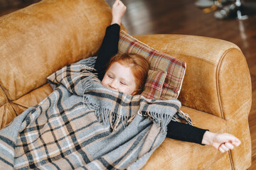 Relaxing child enjoys cozy moment on sofa with blanket in stylish living room during winter...