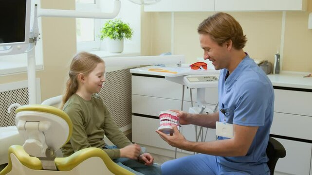 Side view shot of young Caucasian girl playing snap with anatomical tooth model while having fun during consultation in pediatric dentists office, copy space