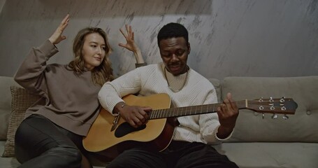 A multiracial couple enjoys quality time together at home as the man plays the acoustic guitar and the woman listens intently. - Powered by Adobe