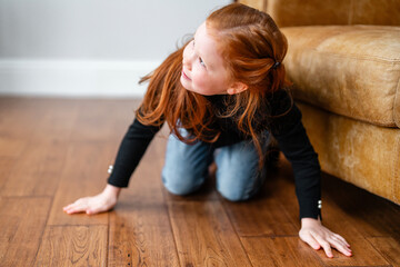 Young girl with red hair playing hide and seek on wooden floor next to couch in cozy living room...