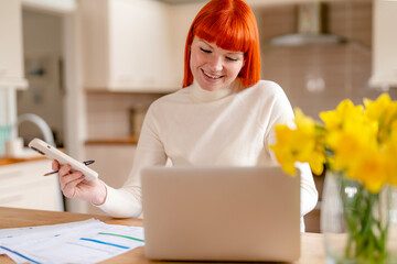 Young woman with red hair works on laptop while holding a smartphone in a bright kitchen,...