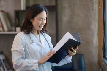 Asian cheerful happy female professional successful doctor practitioner in a white lab coat with a stethoscope standing smiling holding a reading paperwork document in a folder in the hospital.