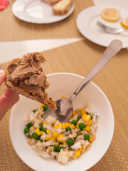 Hand holding slice of bread with pate over bowl of rice, peas, and corn