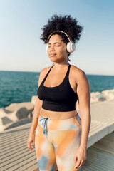Curvy Hispanic woman listening music with headphones by the sea and relaxing after workout outdoors