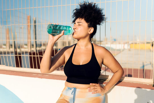 Plus size Hispanic sportswoman drinking water from a bottle after training on a sunny day - Powered by Adobe