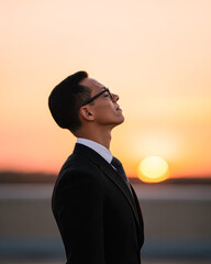 Fototapeta premium A man in a suit and glasses gazes upward thoughtfully against a warm, glowing sunset background.