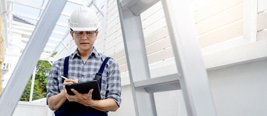 Building inspector man in protective workwear using digital tablet checking for the process of house project. Asian male worker holding smart device at construction site. Shot through the ladder