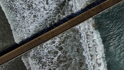 Aerial view of a pier stretching into the turquoise sea, waves crashing against its wooden supports in a frothy dance of white, Pismo Beach, California, United States.