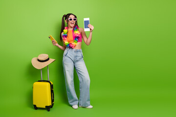 Fashionable young woman holding travel essentials and posing cheerfully against a vibrant green...