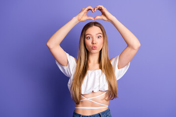 Lovely cheerful woman in stylish white top making heart sign with hands on purple background