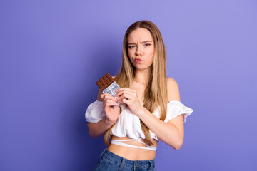 Young fashionable woman holding a chocolate bar posing against a purple wall in casual styling...