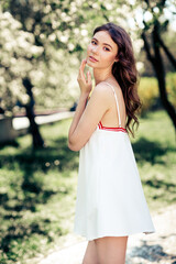 Charming young woman in a white dress posing in a blooming park on a sunny spring day, surrounded...