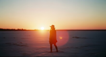 Fototapeta premium Woman in hat standing on a snowy field, facing the setting sun with lens flare.