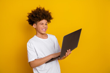 Cheerful teenage boy with curly hair using a laptop against a vibrant yellow background in a casual...