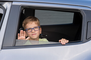 Young boy with glasses waving from car window on a sunny day outdoors. Expresses joy, travel,...