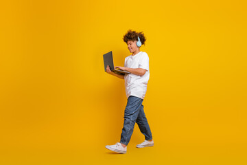 Confident young boy wearing headphones while holding a laptop, standing against a yellow background...