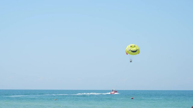 A grinning parasailer soars high above the calm blue ocean, towed by a speedy boat under a clear, sunny sky, enjoying a cheerful water adventure.