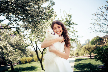 Young woman enjoying a sunny day outdoors with her fluffy white dog in a vibrant blooming park in...