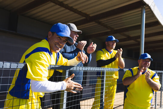 Senior male teammates wearing baseball jerseys leaning on chain-link fence and clapping, copy space