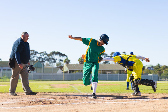 Male runner stepping on home plate as catcher lunging with glove and umpire watching at ballpark