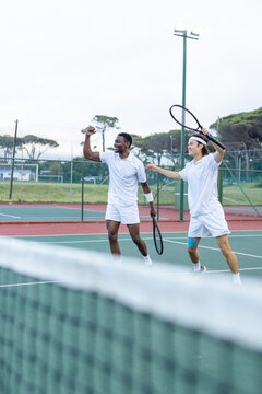 Diverse male friends celebrating winning point while walking on outdoor tennis court with rackets