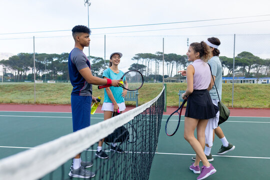 Diverse friends wearing tennis attire pausing at net on outdoor court, exchanging rackets and balls