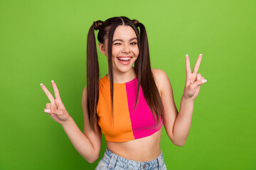 Cheerful young woman winking and gesturing peace signs against a vibrant green background while...