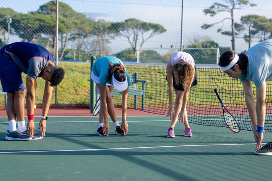 Diverse tennis teammates stretching forward on outdoor court beside net with rackets and bench
