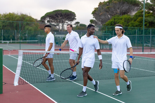 Diverse male tennis players crossing outdoor court net exchanging handshake holding rackets