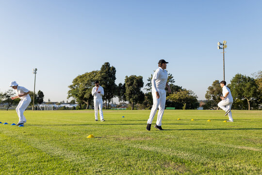 Diverse male players wearing white uniforms fielding on field with cones under floodlights