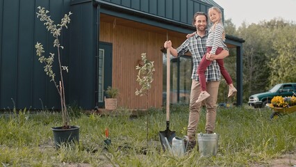 Attractive Caucasian dad holding on his hands little daughter while gardening tree in garden....