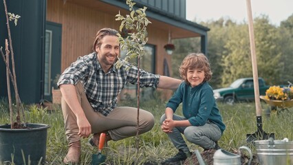 Happy father with his little son sitting near tree and smiling into camera. Positive people having...