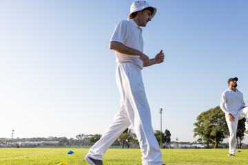 Diverse male teammates walking past cones on cricket field near light pole in white, copy space