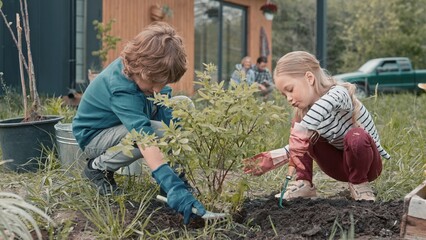 Fototapeta premium Two little children with gloves cultivating ground after planting bush or tree. Obedient kids speaking while working in garden. In blurred background their parents resting and looking at them.