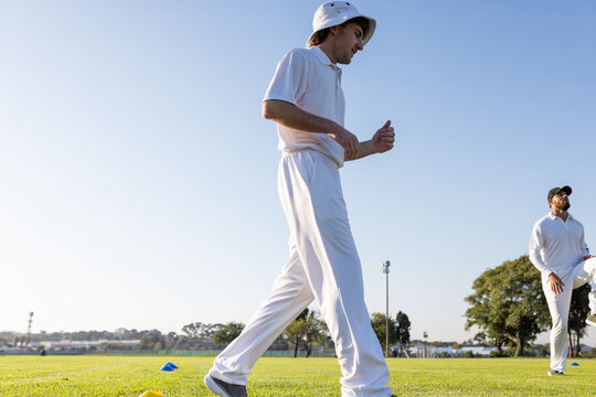 Diverse male teammates walking past cones on cricket field near light pole in white, copy space - Powered by Adobe