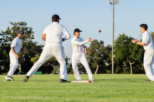 Diverse male teammates standing on grassy field passing red cricket ball by white ground marker - Powered by Adobe
