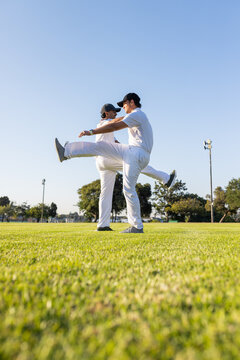 Diverse male teammates stretching forward legs on grass under clear sky wearing white uniforms