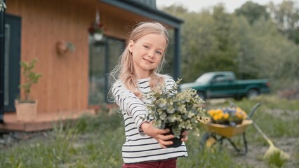 Caucasian little girl holding pot with green plant. Cute blond kid showing on camera beautiful flowerpot. Helping her mother in garden. Having resting day at nature. Concept of childhood.