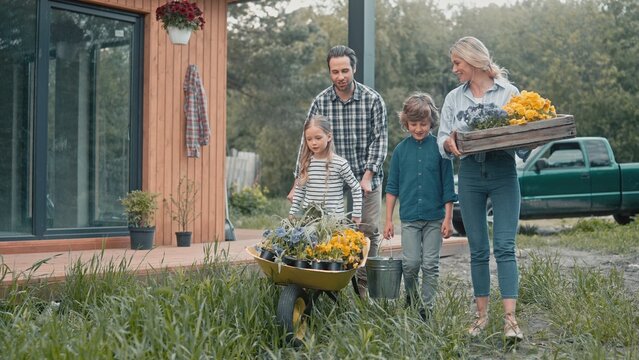 Family with gardening tools walking on green herbs in garden. People going to plant flowers. Father with his daughter riding wheelbarrow full of greenery while his wife and son walking alongside him. - Powered by Adobe